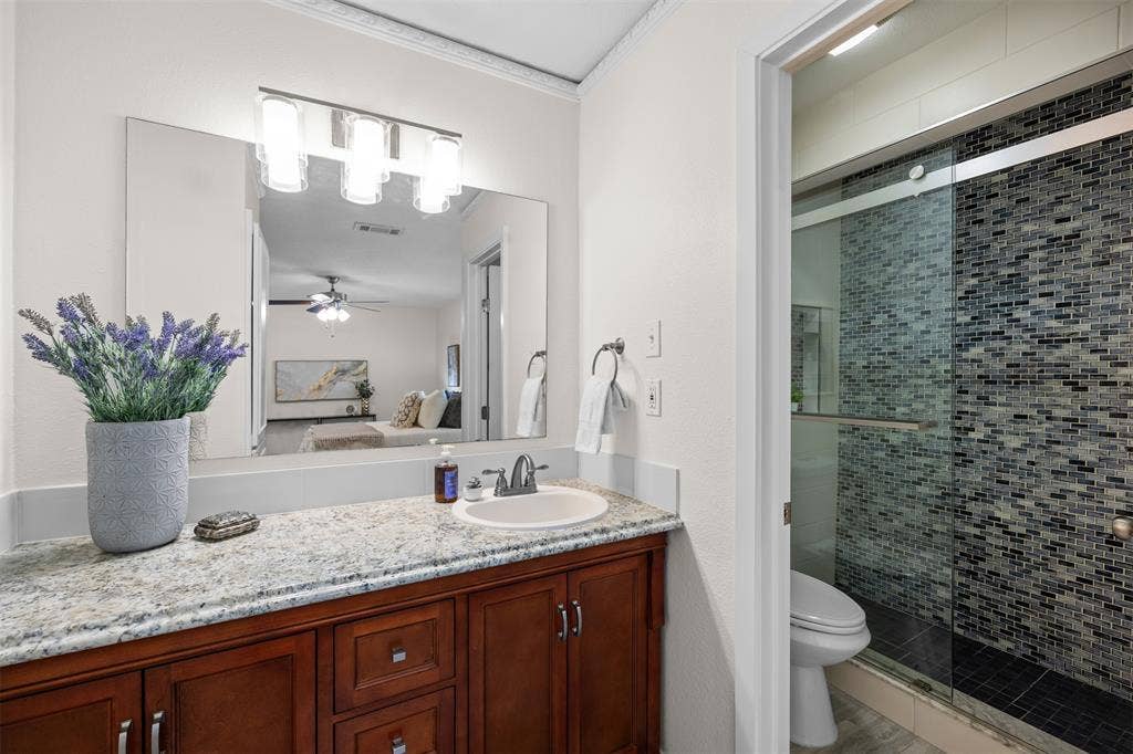 1118 Pacific Drive, Richardson, TX 75081 Granite countertop vanity with an undermount sink, wood-finish cabinetry, a large mirror, and an adjacent shower with glass enclosure and mosaic tile