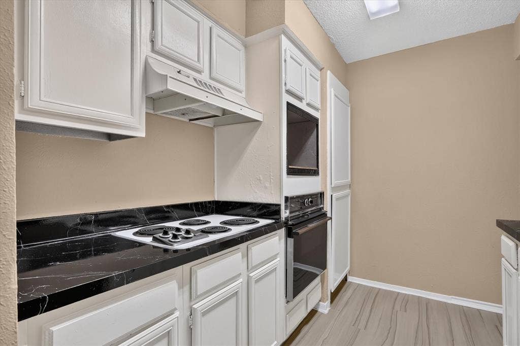 White cabinetry, black countertops, built-in oven, built-in microwave, and wood-finish flooring