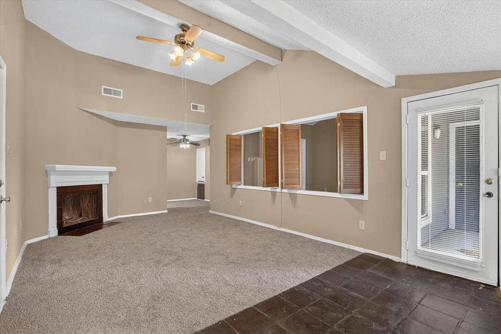 Vaulted ceiling with exposed beams, ceiling fan, fireplace with white mantel, windows with wooden shutters, and a glass-paneled exterior door