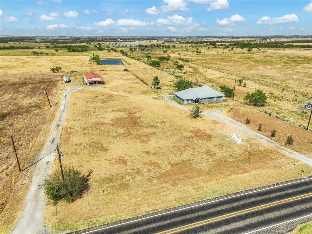 16880 W Fm 455, Celina, TX 75009 Expansive rural property featuring a single-story residence, a separate red-roofed outbuilding, a visible pond, and a paved road frontage