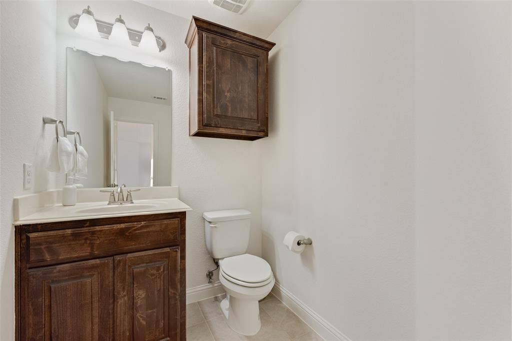 2408 Brisbane Lane, Plano, TX 75075 White toilet, dark wood-finish vanity with an integrated sink, wall-mounted mirror, and overhead tri-light fixture
