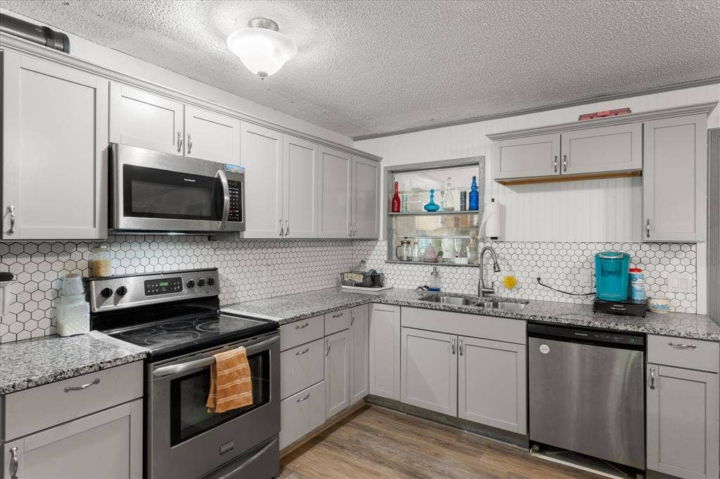 402 E Cook Street, Eustace, TX 75124 Remodeled Kitchen featuring shaker-style cabinetry, stone-finish countertops, stainless steel appliances, a hexagonal tile backsplash, and wood-finish flooring