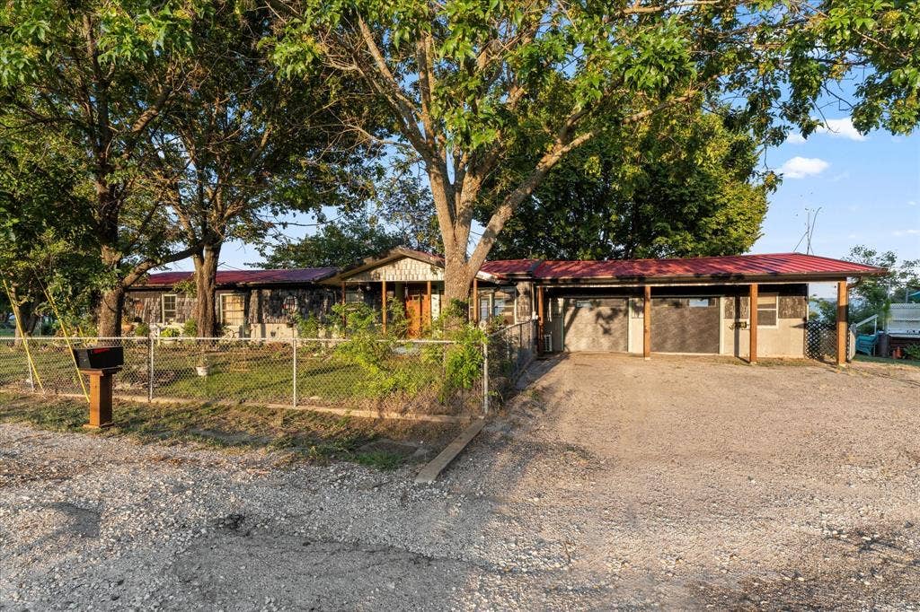 308 Causeway Cove, Seven Points, TX 75143 Red metal roof, gravel driveway, chain link fencing, multiple mature trees, and a covered carport