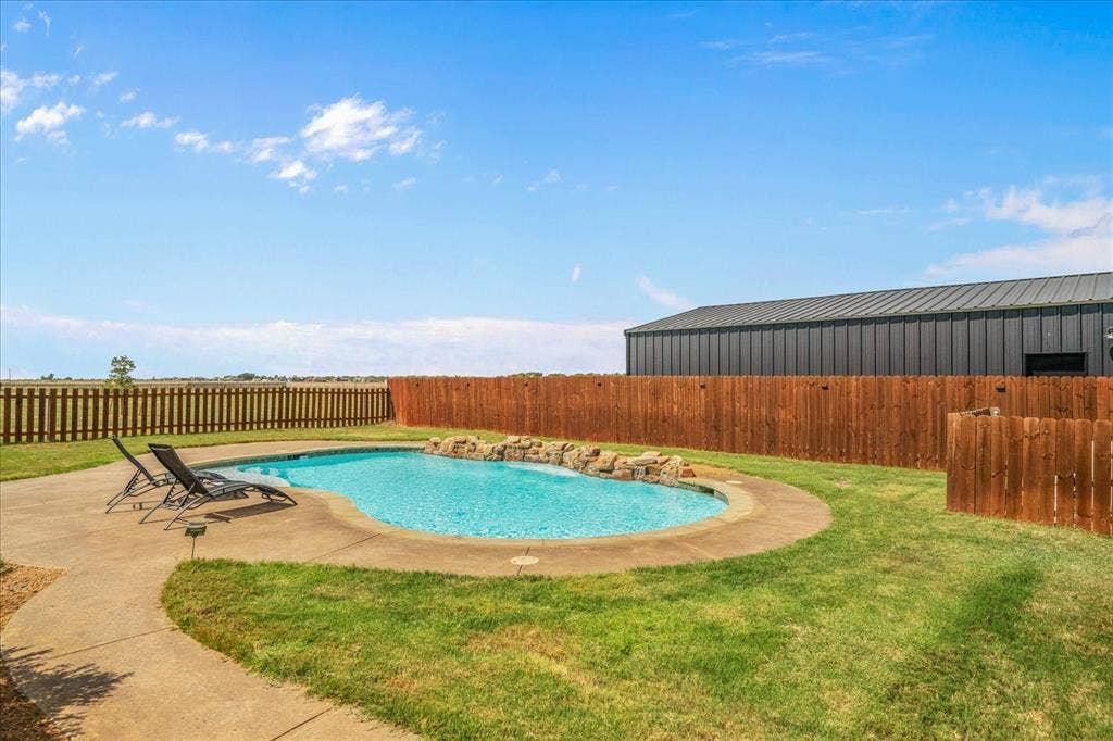 3674 County Road 2142, Caddo Mills, TX 75135 Curved swimming pool with rock detailing, surrounded by a concrete patio, and a grass lawn with wood fencing