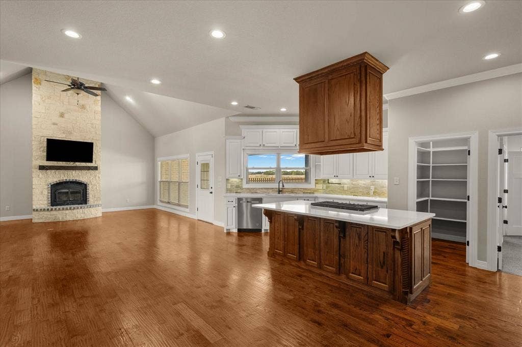 3674 County Road 2142, Caddo Mills, TX 75135 Vaulted ceiling, stone fireplace with a wood-beam mantel, wood-finish flooring, kitchen island with built-in cooktop, and white cabinetry