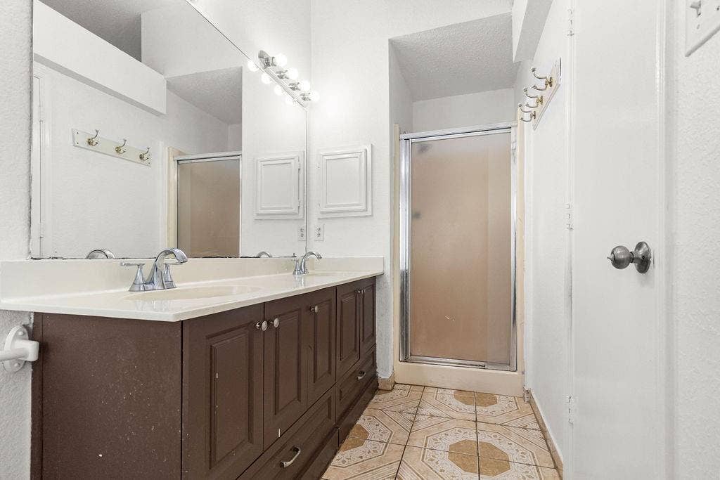 Double-sink vanity with dark-finish cabinetry, solid-surface countertop, full-width wall mirror, and enclosed glass shower stall with patterned tile flooring