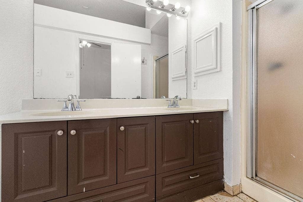 Dual-sink vanity with cabinetry, expansive wall-mounted mirror, overhead vanity lighting, and a glass-enclosed shower stall