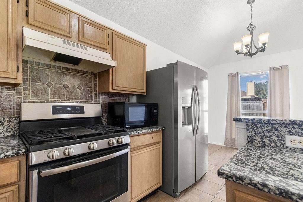 Kitchen featuring wood cabinetry, stone countertops, stainless steel appliances, a tiled backsplash, and a multi-arm chandelier