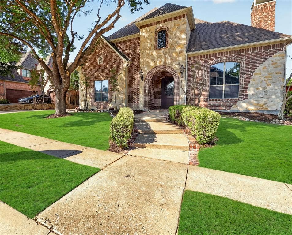 112 Winsley Circle, McKinney, TX 75071 Brick and stone exterior with an arched entryway, mature front tree, and a paved walkway bordered by manicured shrubs