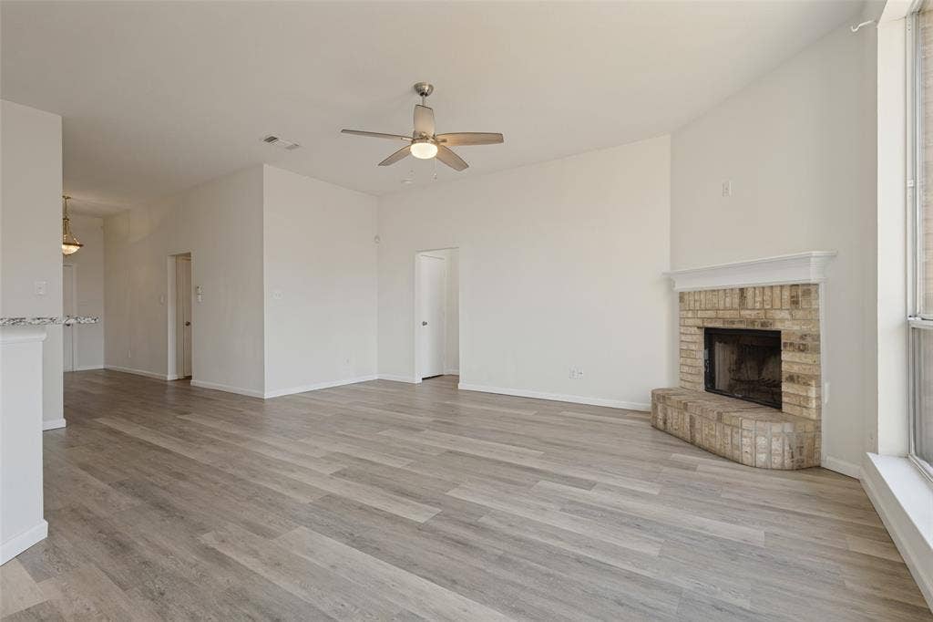 2405 Collier Drive, McKinney, TX 75071 Open-concept living area featuring wood-finish flooring throughout, a ceiling fan with integrated lighting, a brick fireplace with stone surround, and abundant natural light from large windows