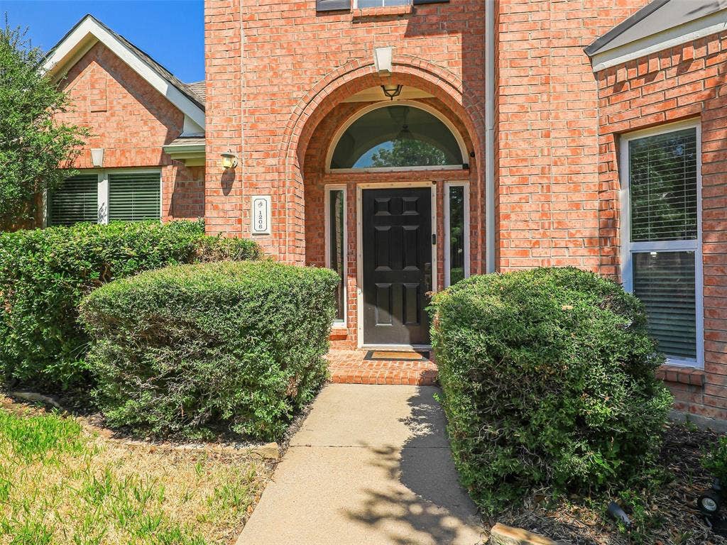1206 Bel Air Drive, Allen, TX 75013 Brick exterior with arched entryway, black paneled door, and established shrubbery