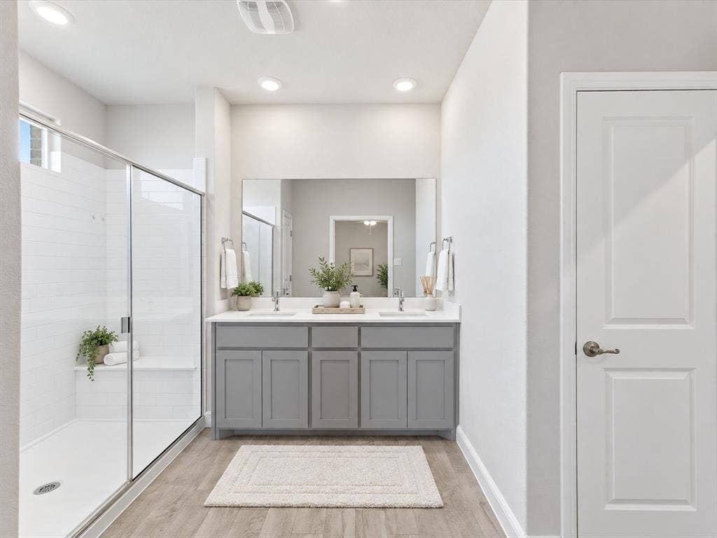 Dual sink vanity with grey cabinetry, rectangular mirror, wood-finish flooring, glass-enclosed shower, and white subway tile surround