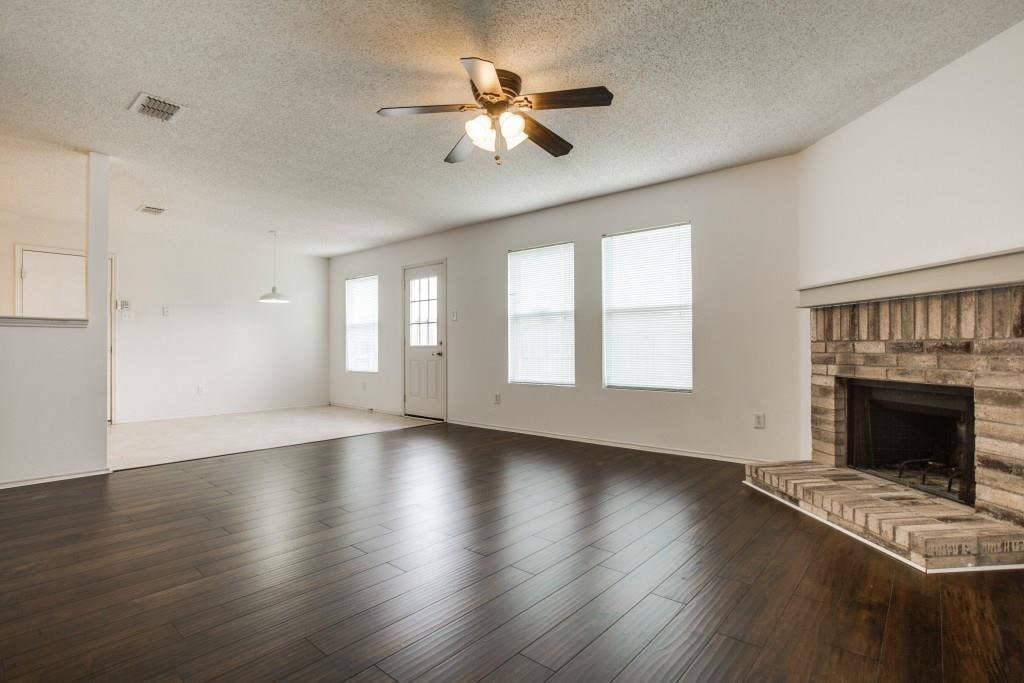 2800 Bissell Way, Wylie, TX 75098 Dark wood-finish flooring, brick fireplace, multiple windows with blinds, ceiling fan with integrated lighting, and a white paneled door with glass inserts