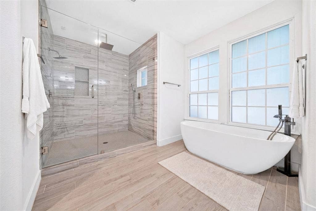 Spacious bathroom featuring a frameless glass shower enclosure, a freestanding soaking tub, wood-finish flooring, and white-framed windows
