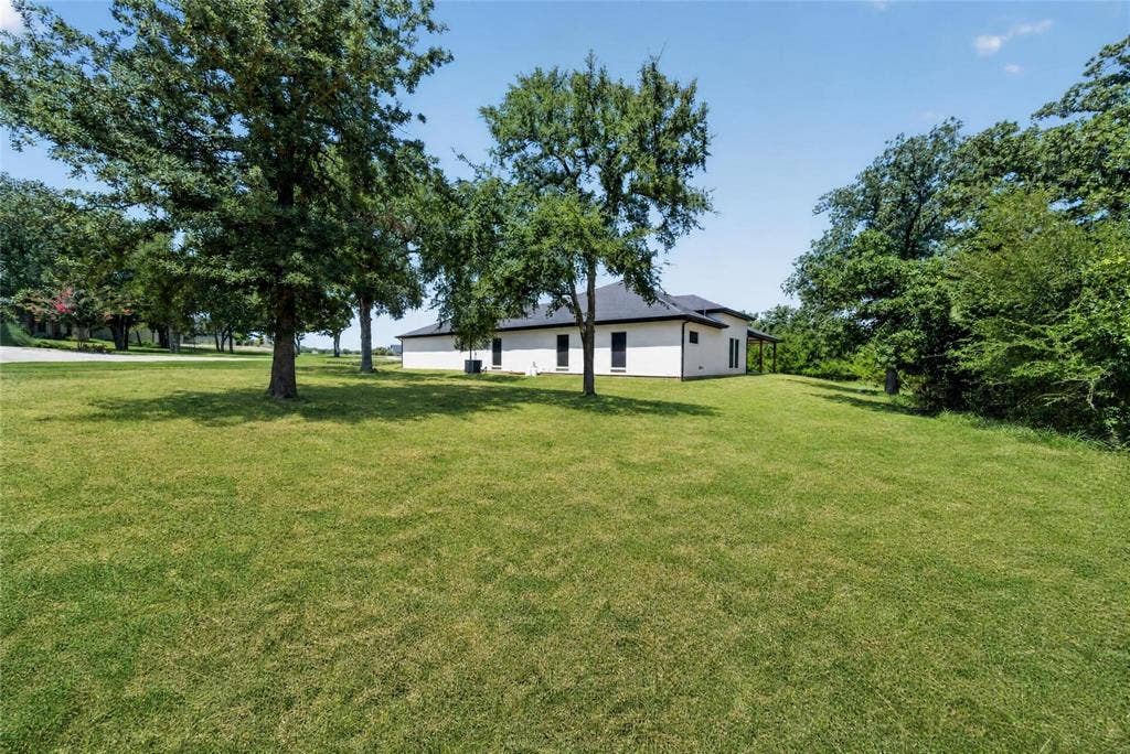 Expansive lawn area with mature trees, a single-story residence with a dark roof, and a covered porch