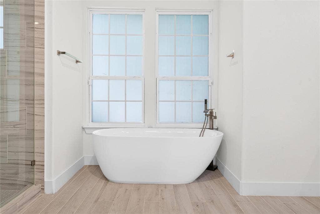 Freestanding soaking tub with floor-mounted faucet, wood-finish flooring, dual pane windows, and a glass-enclosed shower with stone-look tile