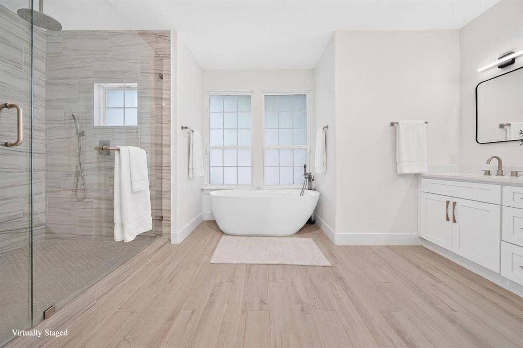 Expansive bathroom featuring a freestanding soaking tub, glass-enclosed shower with tile surround, wood-finish flooring, dual-sink vanity with white cabinetry, and modern lighting fixture