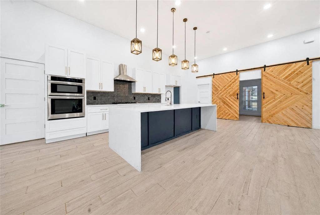 Kitchen featuring white cabinetry, stainless steel appliances, a dark tile backsplash, a large center island with a sink, and light wood-finish flooring