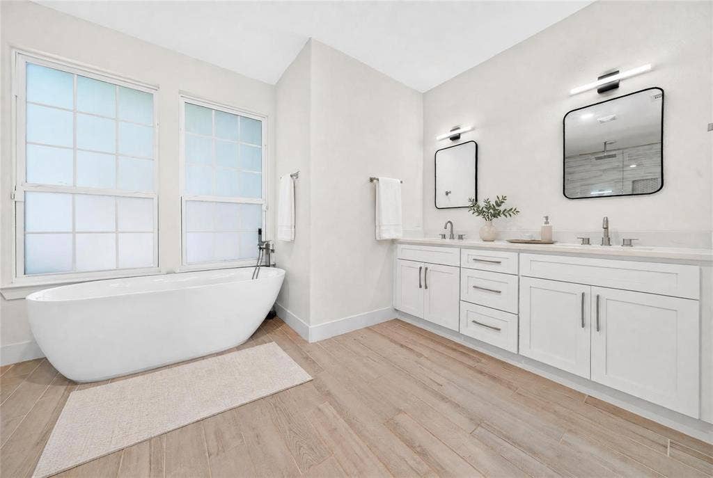 Freestanding soaking tub, dual windows with privacy glass, wood-finish flooring, dual vanity with white cabinetry, and wall-mounted light fixtures