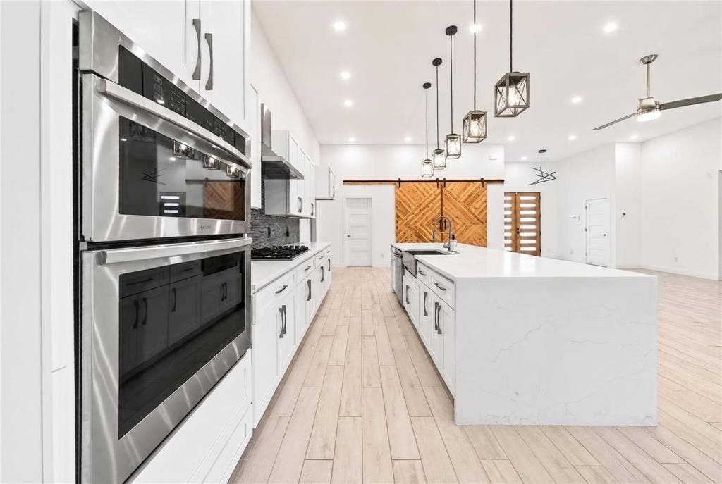 Spacious kitchen featuring an expansive center island, stainless steel double ovens, white cabinetry with dark hardware, wood-finish flooring, and pendant lighting