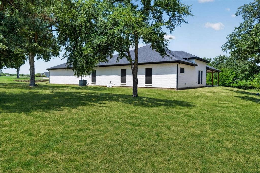 Expansive lawn with mature trees, white brick exterior, dark-framed windows, and a covered patio area