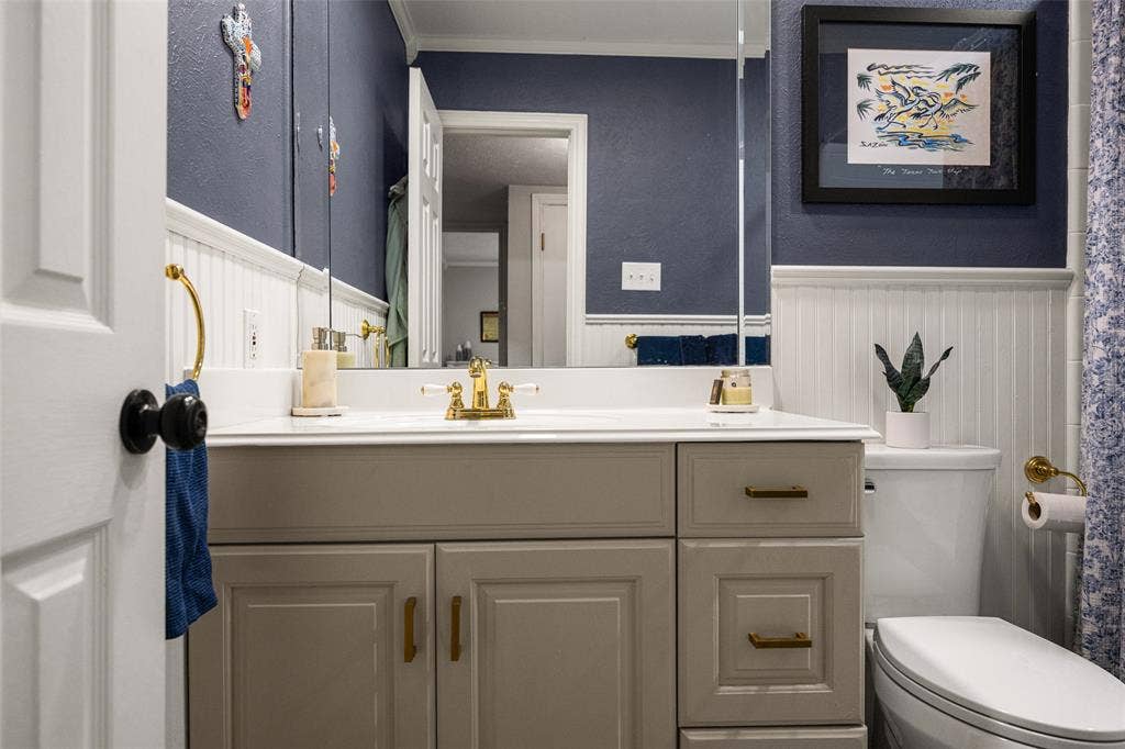 10945 Middle Knoll Drive, Dallas, TX 75238 Vanity with integrated sink, gold-tone fixtures, wall-mounted mirror, and beadboard wainscoting