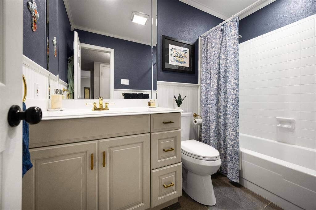 10945 Middle Knoll Drive, Dallas, TX 75238 Vanity with gold-tone fixtures, wood-look flooring, white paneled wall detailing, and a white tiled shower surround