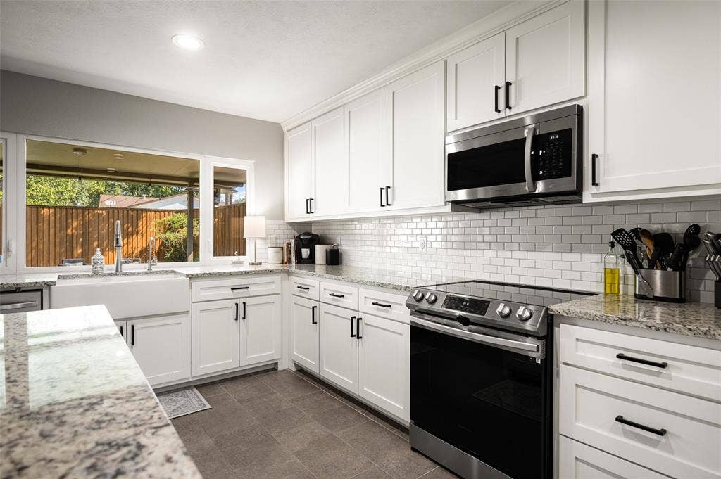 10945 Middle Knoll Drive, Dallas, TX 75238 White shaker cabinetry with matte black hardware, stainless steel appliances, subway tile backsplash, light-toned stone countertops, and an undermount farmhouse sink