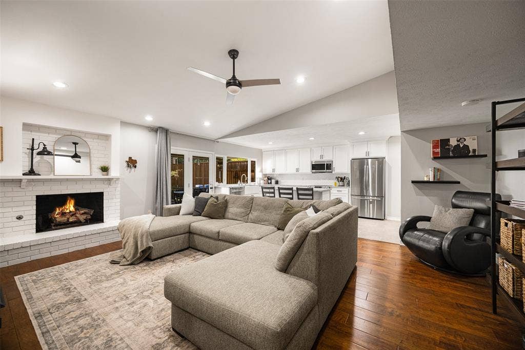 10945 Middle Knoll Drive, Dallas, TX 75238 Open-concept living space featuring wood-finish flooring, a white brick fireplace, vaulted ceilings, recessed lighting, and a modern ceiling fan