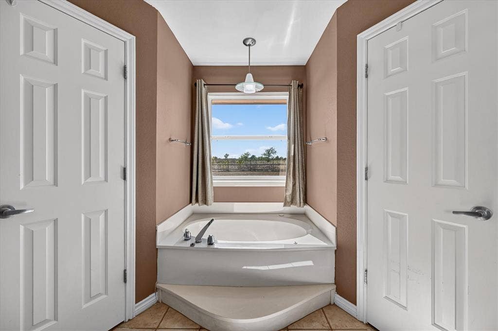 Built-in soaking tub with a surrounding white countertop, under a window with drapes, complemented by a suspended light fixture