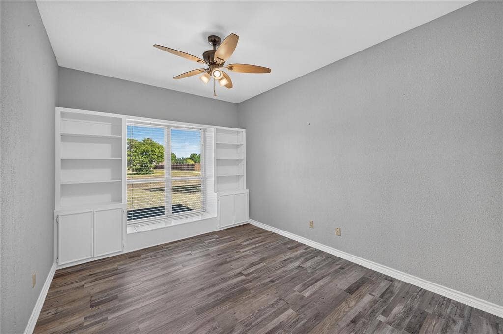 Wood-finish flooring, built-in shelving, window with blinds, ceiling fan with light fixture