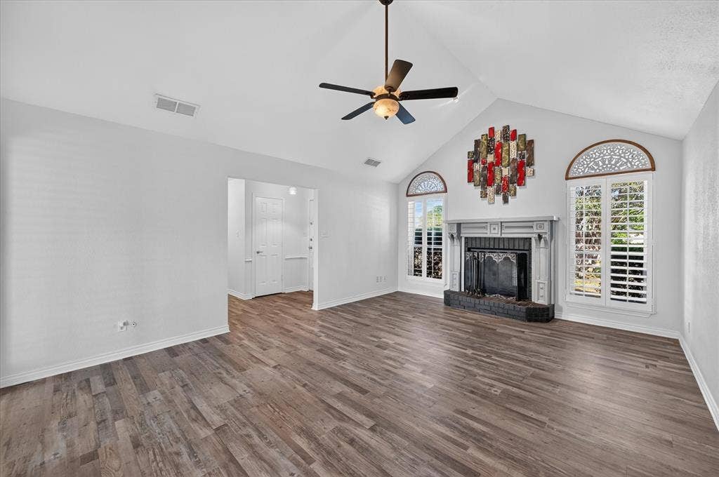 Vaulted ceiling with a ceiling fan, fireplace with a mantel, wood-finish flooring, and arched windows with plantation shutters