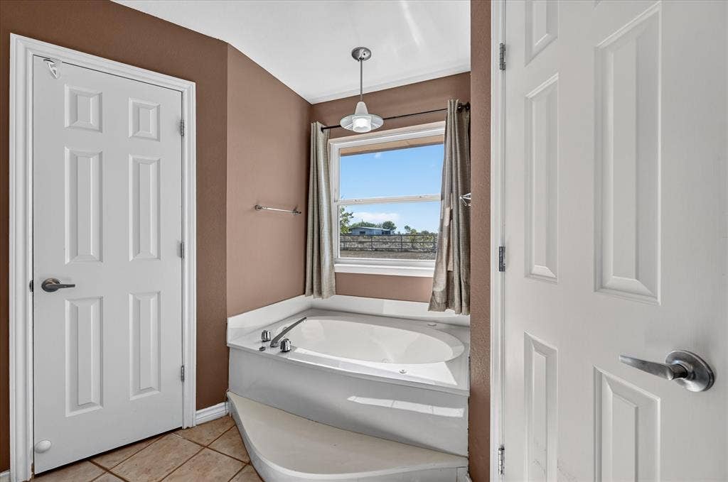 Built-in corner bathtub with a tiled surround, overhead lighting fixture, and window