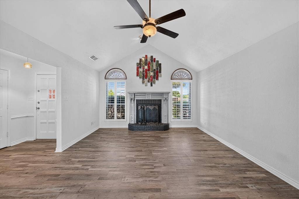 Vaulted ceiling with a ceiling fan, fireplace with a stone hearth, arched windows with plantation shutters, and wood-finish flooring