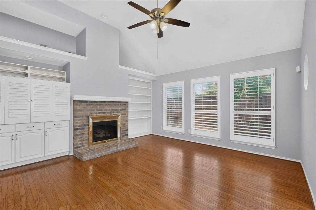 2702 Garden Springs Drive, Richardson, TX 75082 Spacious great room featuring warm wood-finish flooring, a brick fireplace with built-in surround, white shutter cabinetry with integrated storage, multiple glass doors with integrated blinds flooding the space with natural light, and a brass ceiling fan