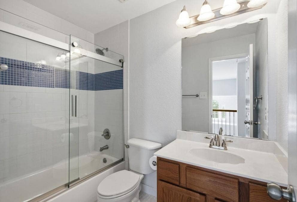 Tub/shower combination with sliding glass doors and blue mosaic tile accent, wood-finish vanity with white countertop, and wall-mounted mirror with overhead light fixture