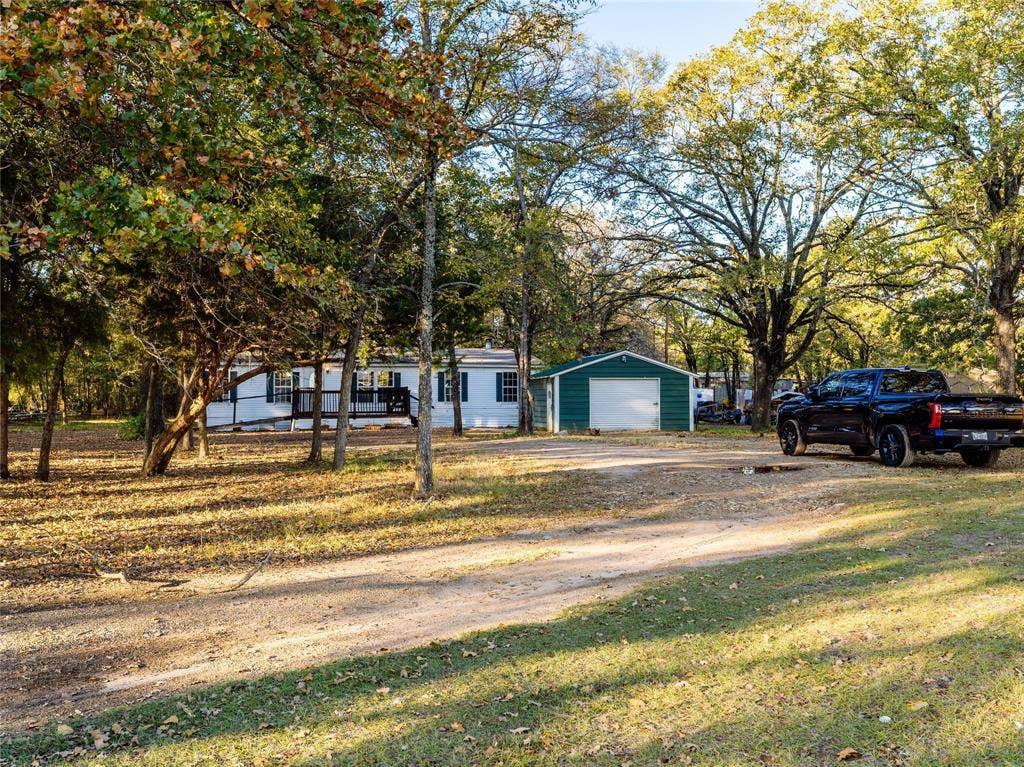 Expansive lot with mature trees, a white-sided residence, a covered porch, and a detached green storage structure