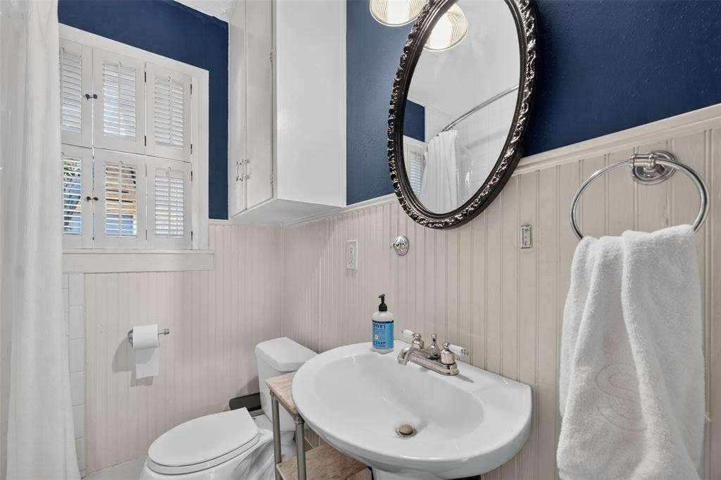 5630 Vanderbilt Avenue, Dallas, TX 75206 Pedestal sink with chrome fixtures, beadboard wainscoting, wall-mounted cabinetry, window with plantation shutters, and an oval vanity mirror