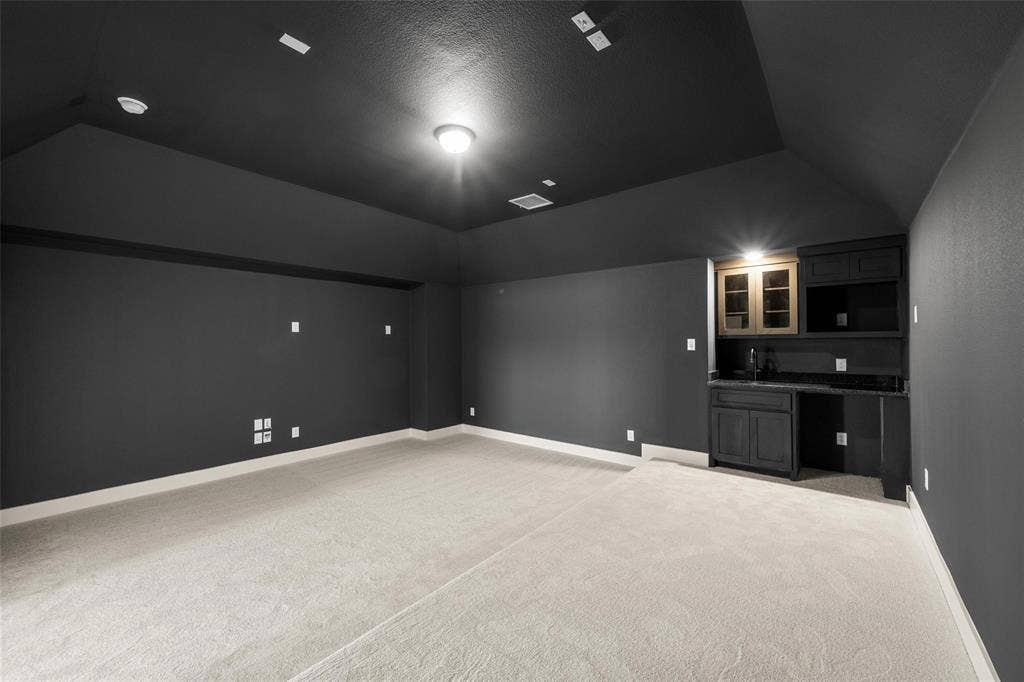 Media Room Vaulted ceiling, neutral wall-to-wall carpeting, and a built-in wet bar featuring dark cabinetry, stone countertops, and upper glass-front display storage