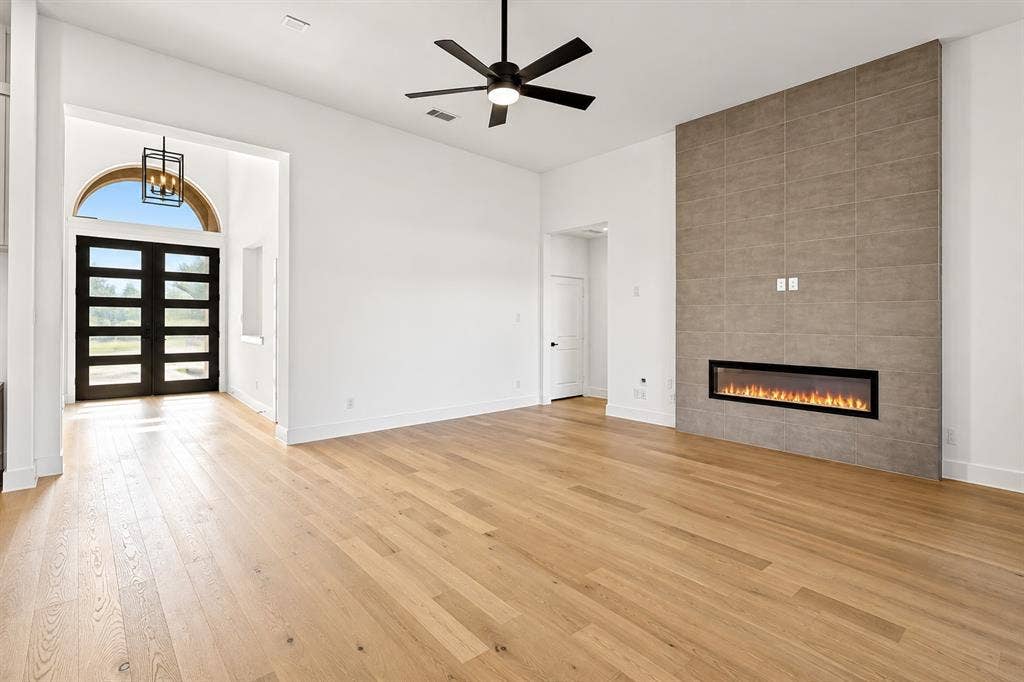 High-ceiling living area featuring wood-finish flooring, a floor-to-ceiling tiled fireplace surround with a linear electric insert, a black multi-blade ceiling fan, and double front doors with a decorative arched transom window