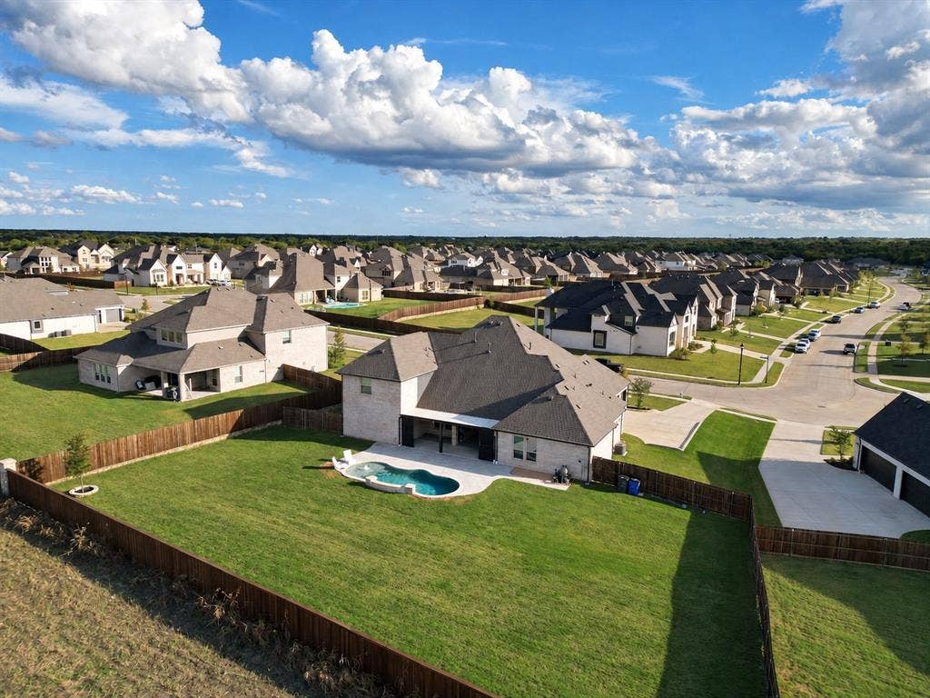 Aerial perspective of a residential property featuring an in-ground pool, covered patio, wood fencing, and expansive lawn