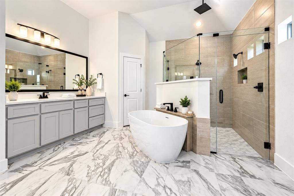 Double vanity with gray shaker-style cabinetry, freestanding soaking tub, glass-enclosed walk-in shower with tiled surround, and large-format stone-finish flooring