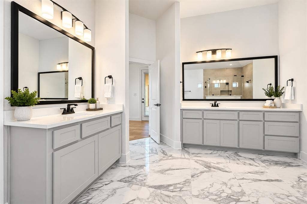 Dual vanities featuring grey shaker-style cabinetry, white solid-surface countertops, black framed mirrors, and light-tone large-format stone-patterned floor tile