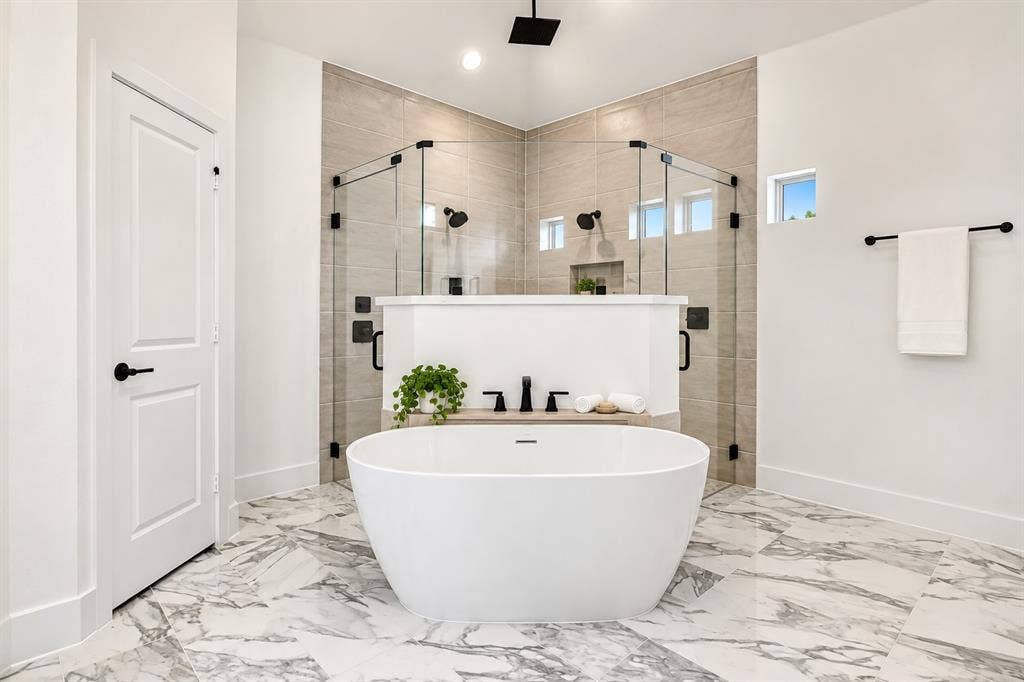 Freestanding soaking tub, glass-enclosed walk-in shower with dual showerheads, stone-tile flooring, and matte black hardware