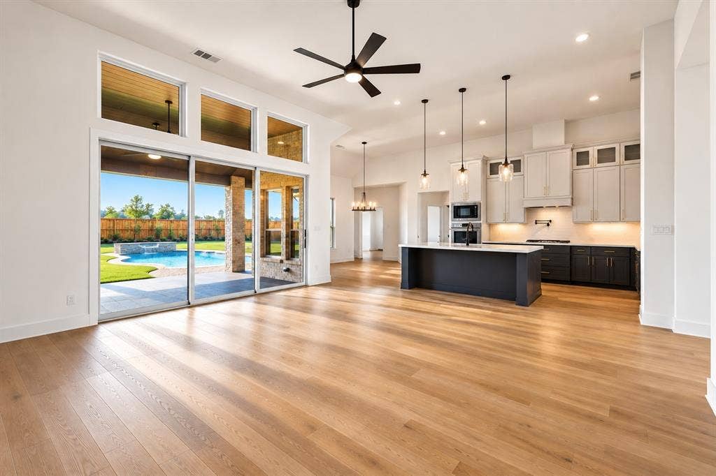 Expansive wood-finish flooring, a modern kitchen with a large island, stainless steel appliances, and abundant natural light from large sliding glass doors, leading to a visible outdoor pool area
