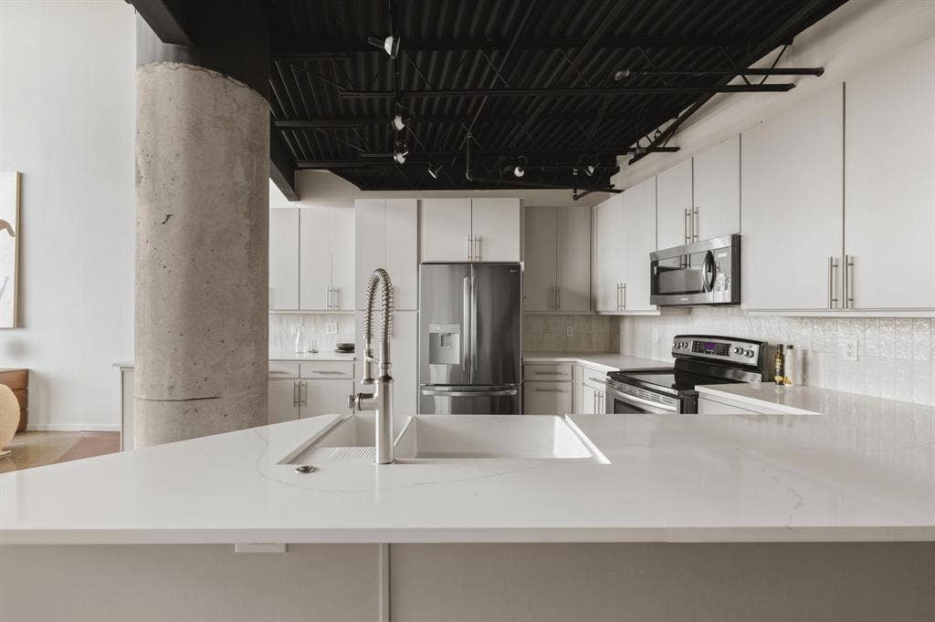 Modern kitchen featuring white cabinetry, stainless steel appliances, a white basin sink with a chrome faucet, and exposed concrete column