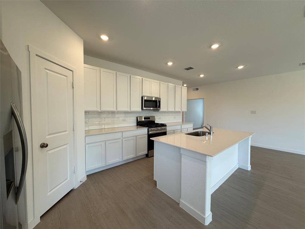 Open-concept kitchen featuring white cabinetry, stainless steel appliances, a center island with an integrated sink, recessed lighting, and wood-finish flooring