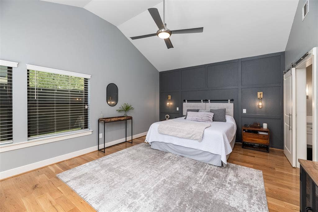 Vaulted ceiling with a ceiling fan, accent wall with paneling, wood-finish flooring, two windows with horizontal blinds, and a barn door