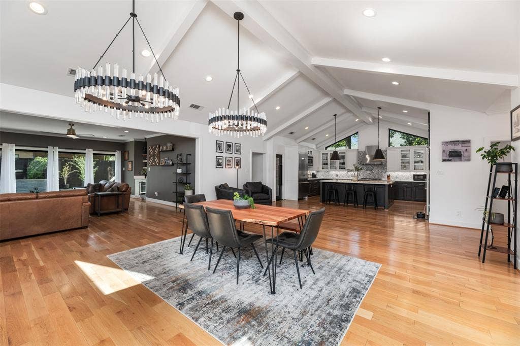 Vaulted ceilings with exposed beams, wood-finish flooring, two contemporary chandeliers, modern kitchen with island seating, and large windows
