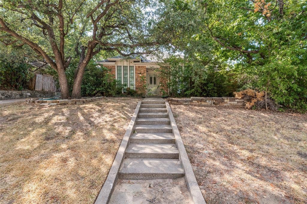 1901 Wedgewood Drive, Grand Prairie, TX 75050 Brick facade residence with a concrete walkway, mature trees, and a stone retaining wall